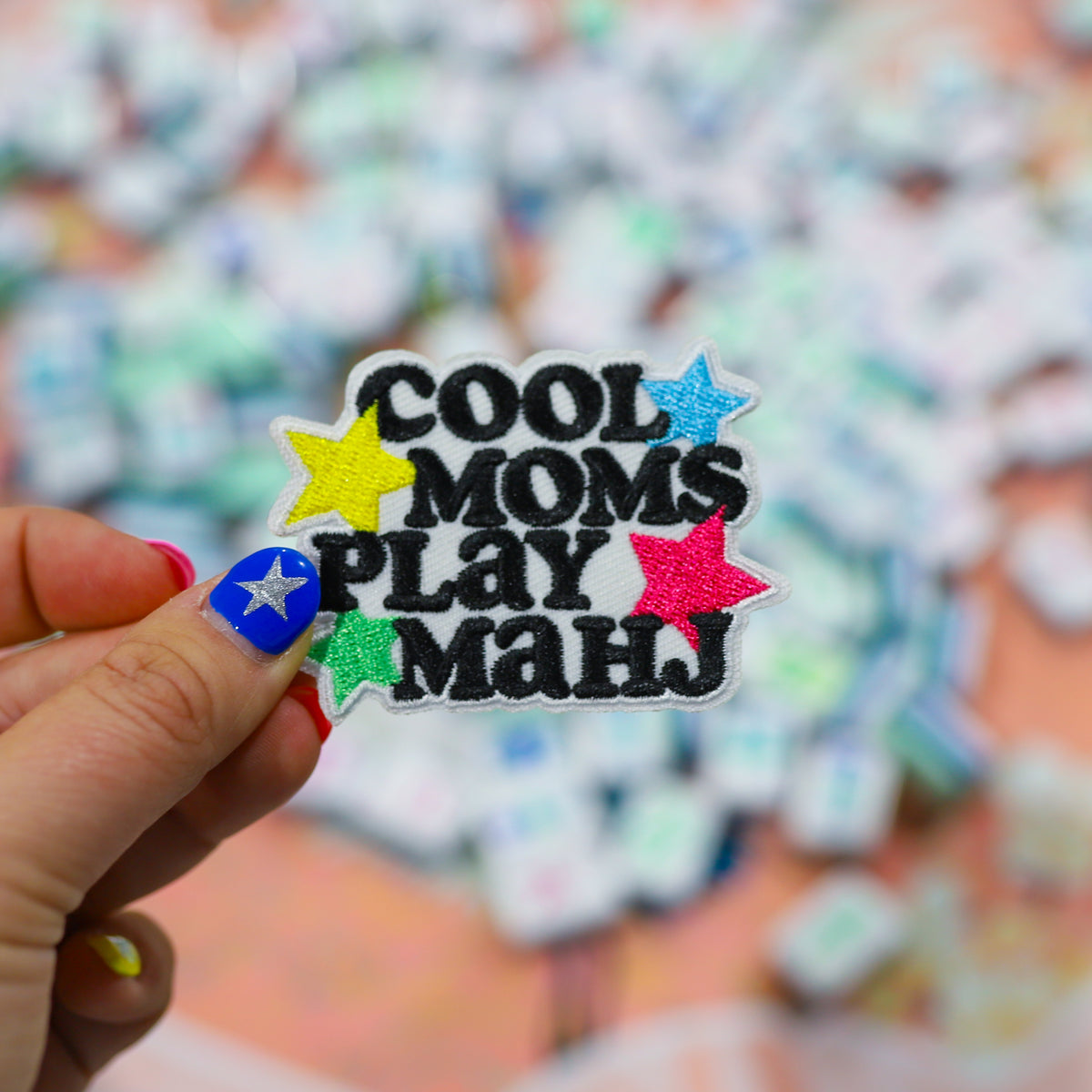 A hand with pink and blue nail art holds a glittery embroidered patch that reads 'COOL MOMS PLAY MANJ' in black letters with colorful star accents. The background is a soft-focus pile of similar patches.