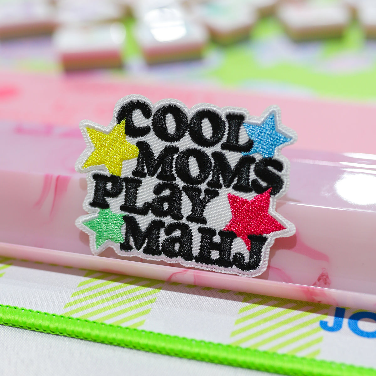 An embroidered patch with the text 'COOL MOMS PLAY MAHJ' in black letters, surrounded by five colorful glittery stars in yellow, blue, green, and red, with a white border. The patch is resting on a pink translucent pen or stylus over a patterned desk.